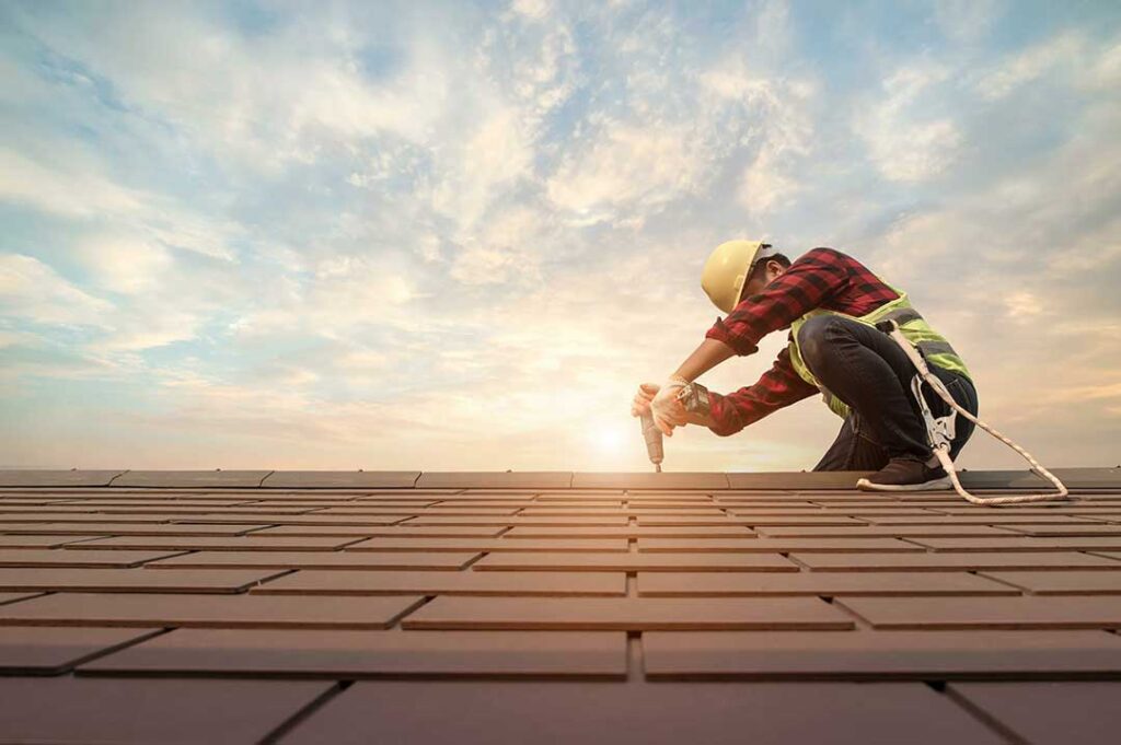 Workers install layered roofing materials on a pitched house roof beside ladders and protective ground coverings.